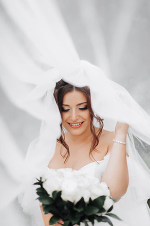 Curly brunette bride in a white dress, covered with a veil, poses for the camera with a bouquet of...