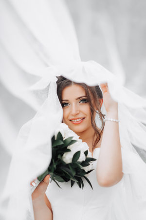Curly brunette bride in a white dress, covered with a veil, poses for the camera with a bouquet of roses. Portrait of the bride. Beautiful makeup and hair. Wedding in natureの写真素材