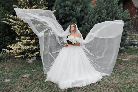 A curly-haired brunette bride in a white dress and veil, flowing beautifully in the air, stands in...