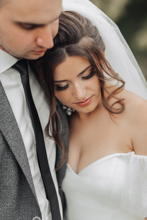 Portrait of the bride and groom in nature. The groom and the brunette bride in a white long dress are hugging and looking down. Stylish groom. Beautiful hair and makeupの写真素材