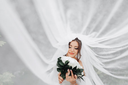 Curly brunette bride in a white dress, covered with a veil, poses for the camera with a bouquet of roses. Portrait of the bride. Beautiful makeup and hair. Wedding in natureの写真素材