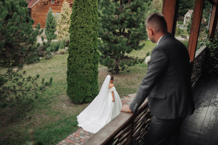 Wedding photo. The bride in a voluminous white dress and a long veil walks in the garden along a stone path, the groom stands on the balcony and looks at her. View from above. Beautiful makeupの写真素材