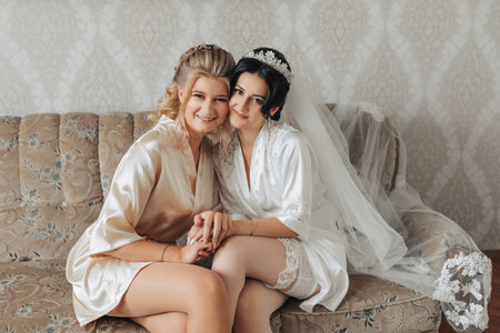 A brunette bride and her friend are sitting on a sofa in the bride's room, dressed in robes, smiling and posing. Wedding portrait wedding photo in a light key. Cropped photo. Morning of the bride. A gorgeous crownの写真素材
