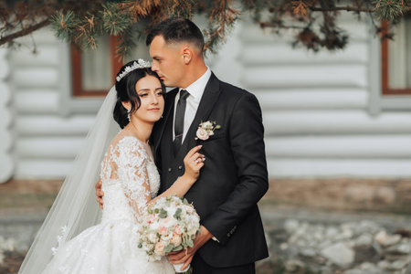 Wedding portrait in nature. The brunette bride and groom in a white long dress are standing and hugging against the background of coniferous trees and a white hut. Stylish groom. Beautiful hair and makeupの写真素材