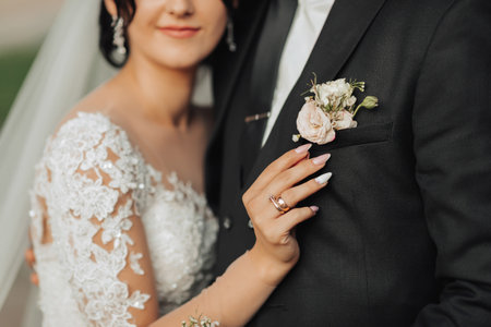 Wedding portrait in nature. Cropped photo. Accent on the boutonniere. The bride and groom, a brunette in a white long dress, stand hugging, the bride touches the groom. Stylish groom.の写真素材