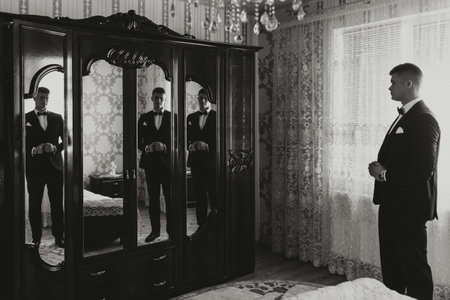 black and white photo. A handsome man in a black suit poses in front of the camera, reflected in the...