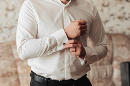 Photo details. A man fastens a button on the sleeve of a white shirt. Front view. Daylight. Fashion...