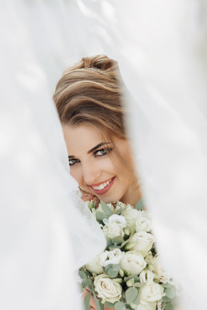 Curly blonde bride in a white dress, covered with a veil, poses for the camera with a bouquet of roses. Portrait of the bride. Beautiful makeup and hair. Wedding in natureの写真素材