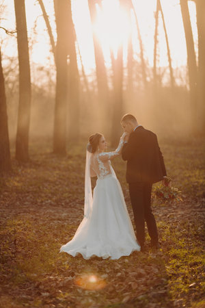 Wedding photo. The bride and groom are walking in the forest. The groom holds a bouquet and kisses the hand of his beloved. Long wedding dress. Couple in love. Autumn sunlight.の写真素材