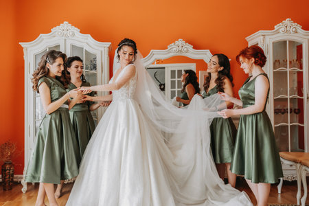 Enjoying the preparations for the wedding. bridesmaids helping the bride to put on the veil and dress and smiling while standing in the roomの写真素材