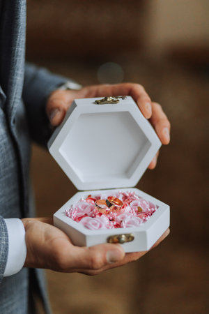 Wedding gold rings on a decorative white box with pink flowers, lying in the hands of the groom,...