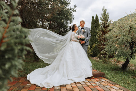 Full length side view of one beautiful sensual young brunette bride in long white wedding dress and veil standing in park holding bouquet together with groom outdoors on natural background, horizontalの写真素材