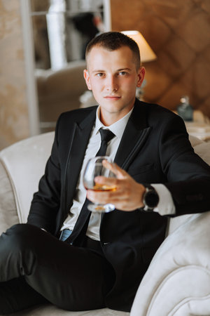 a young man sitting in a chair with a glass of alcoholic drink in a hotel room, a young businessman wearing a black suit and a white shirt with a tie. The groom is preparing for the wedding ceremony.の写真素材