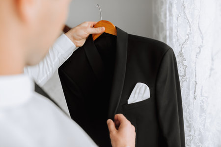 a young man holds his black jacket on a hanger in his hands. The groom is preparing for the wedding ceremony. Detailed close-up photo of handsの写真素材