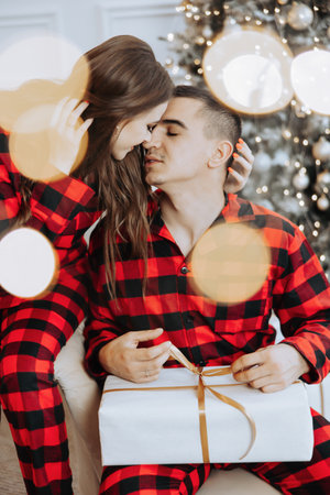 Young beautiful family couple in identical red pajamas near the Christmas tree rejoice and exchange Christmas gifts in their hands. New Year's holidays and gifts under the Christmas treeの写真素材