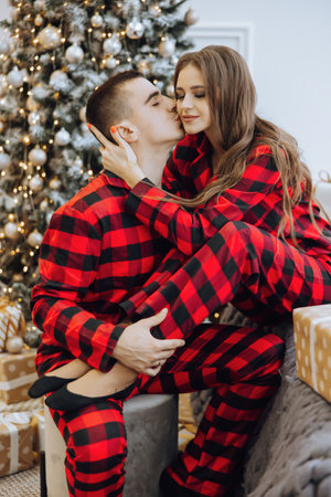 A young beautiful married couple in the same red pajamas, near the Christmas tree, are smiling, hugging and kissing. New Year holidays and gifts under the Christmas treeの写真素材