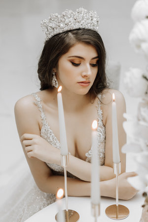 Glamorous portrait of a girl at a table with burning candles on candlesticks, a bride in a wedding dress and a crown on her head. Vertical photoの写真素材