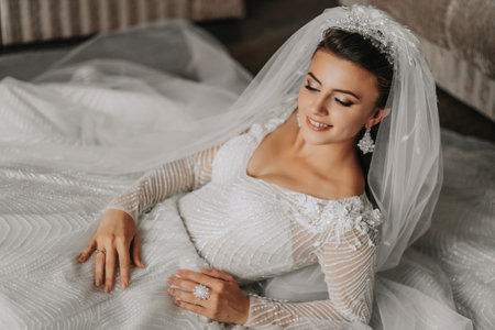 A brunette bride, dressed in a lace dress, poses while lying on the floor. Beautiful hair and make-up, open bust. Wedding portrait. Gorgeous silver jewelryの写真素材