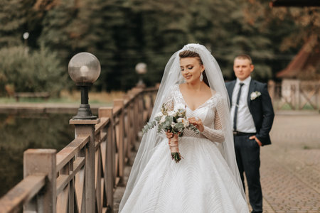 Wedding portrait. A brunette bride in a long dress holds her bouquet of roses, peonies and greenery, the groom in a classic suit walks behind her on the bridge.の写真素材