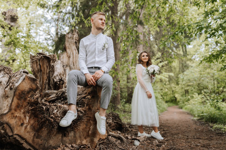 Wedding walk in the forest. The bridegroom is seated on a felled old tree in the foreground. Country wedding concept.の写真素材