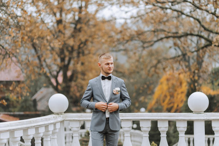 The groom in a classic suit stands by the white railing against the background of tall treesの写真素材