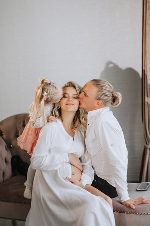 studio portrait of a beautiful pregnant young woman in a white dress with a train sitting on a sofa with her little daughter and her husbandの写真素材