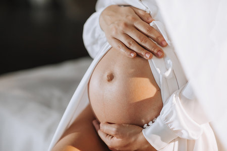 studio portrait, close-up, of a beautiful pregnant young woman on a white bed, holding her pregnant belly with her handsの写真素材