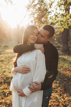 Happy young parents, mom and dad, hugging their pregnant belly, enjoying a beautiful moment at sunset. Stylish pregnant couple holding hands on belly and hugging in sunlightの写真素材