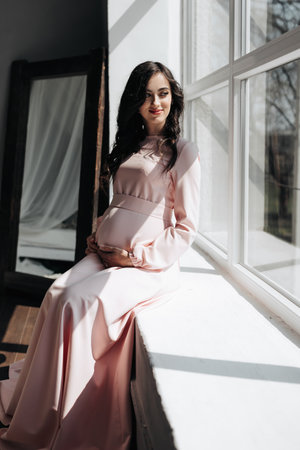 A beautiful pregnant woman in a pink dress holds her hands on her stomach. A loving mother is waiting for the birth of a child. Concept of pregnancy, motherhood, preparation and waiting.の写真素材