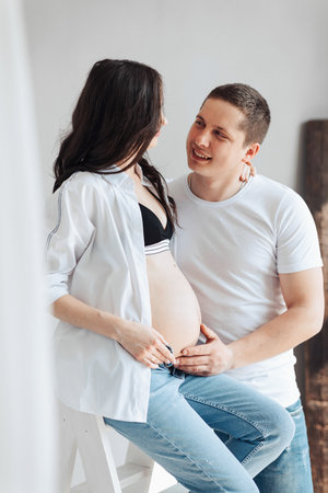 modern beautiful pregnant woman in jeans and shirt with man hugging her tummy with hands in beautiful sunlight. Concept of pregnancy, motherhood, preparation and waiting.の写真素材
