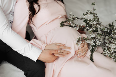 Close up photo of happy pregnant woman at home with man holding her belly and wearing white shirtの写真素材