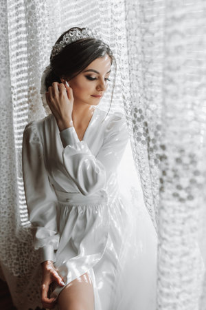 portrait of a beautiful girl in a robe with a crown on her head. Fashion, glamour, concepts. The morning of the bride, the bride in a robe near the window in natural lightの写真素材
