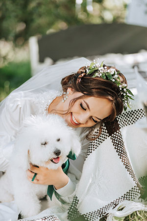 young elegant bride in a white dress with a tiara of fresh flowers on her head posing in the garden with her poodle dogの写真素材