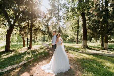 Fashionable groom and cute bride in white dress with tiara of fresh flowers, hugging, laughing in park, garden, forest outdoors. Wedding photography, portrait of smiling newlyweds. Wide angleの写真素材