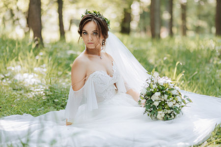 a beautiful bride in a white dress with a wreath of fresh flowers is sitting on the green grassの写真素材