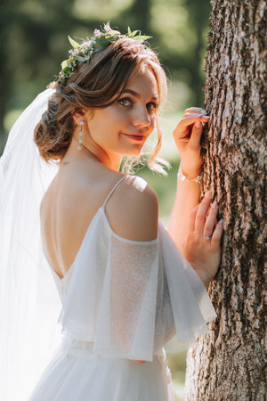Portrait of a beautiful cute bride girl in an elegant white dress posing near a tree in the forest on a sunny summer dayの写真素材