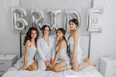 Young bridesmaids in white silk dresses on a bed in the bride's room. Beautiful women celebrating bachelorette party sitting on bed.の写真素材