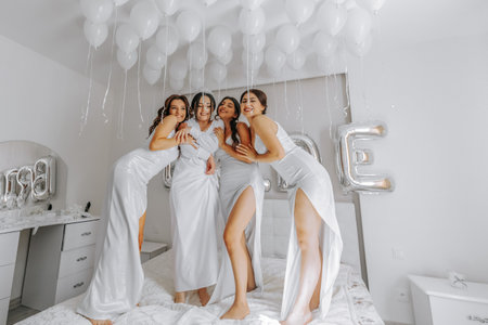 Young bridesmaids in white silk dresses on a bed in the bride's room. Beautiful women celebrating bachelorette party sitting on bed.の写真素材