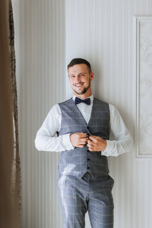 A stylish groom with a beard is preparing for the wedding ceremony. Groom's morning. A businessman fastens a gray vest. The groom is getting ready in the morning before the wedding ceremonyの写真素材
