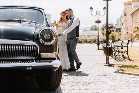 Front view of married bride and groom wearing festive clothes walking through the city to their retro car, holding wedding bouquet, smiling on wedding dayの写真素材