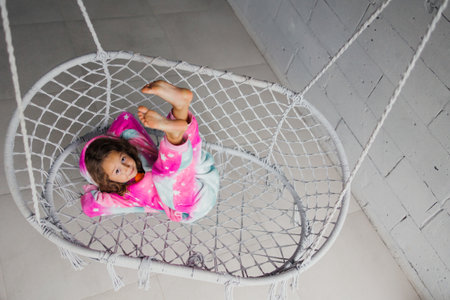 A happy little girl in pink pajamas is lying on a wicker swing and playing with her legs up on the porch.の写真素材