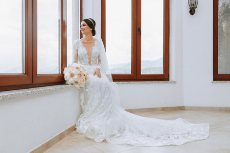 the bride in an evening dress sits on the windowsill against the background of large windows. Beautiful young bride. Free spaceの写真素材
