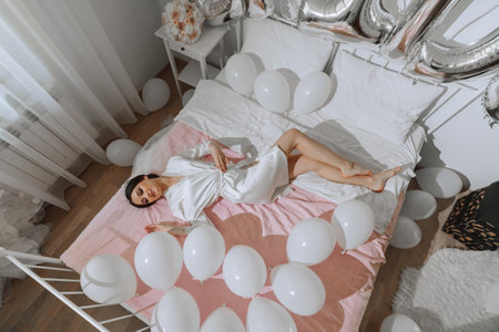 A brunette bride in a white robe and veil lies on her bed near white balls. Wedding concept. Morning of the brideの写真素材