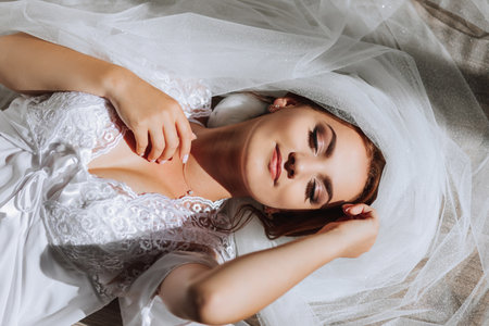 A beautiful young bride in a white robe poses for a photographer, lying on the floor, on a veil. Wedding photography, close-up portrait, chic hairstyle.の写真素材