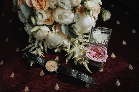 Photo of wedding accessories. Luxurious bouquet of the groom. Gold rings on a stand with flowers, a black classic watch on a burgundy background.の写真素材