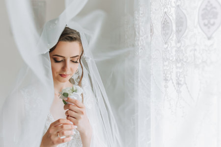 The bride is dressed in an elegant dress, covered with a veil, posing and holding a boutonniere. Wedding photo, morning of the brideの写真素材