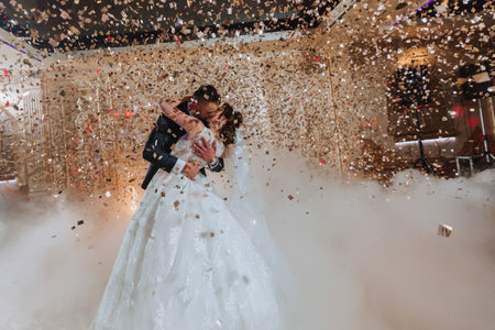 Gorgeous bride and stylish groom dancing under golden confetti at wedding reception. Happy wedding couple performing first dance in restaurant. Romantic momentsの写真素材