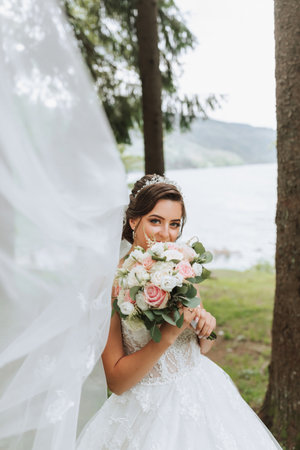 Portrait of a bride with a bouquet against the background of tall trees and a lake. A beautiful young bride is holding a wedding bouquet in her hands, the bride's veil is blowing in the wind.の写真素材