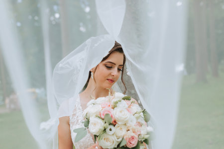 the bride in a wedding dress with a long train and a veil holds a wedding bouquet of roses, under the veilの写真素材