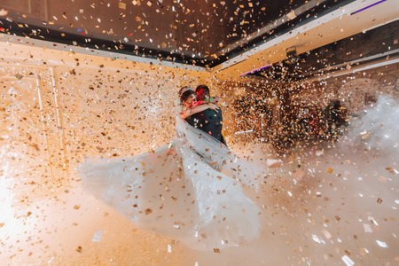 Gorgeous bride and stylish groom dancing under golden confetti at wedding reception. Happy wedding couple performing first dance in restaurant. Romantic momentsの写真素材